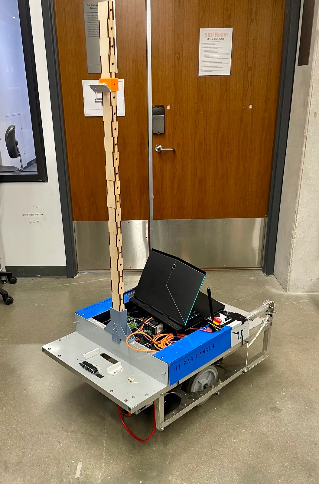 The UT RAS Ranger: an aluminum-framed ground vehicle with a laptop, wiring, a forward-facing sensor, and a tall mast carrying an overhead camera mount.